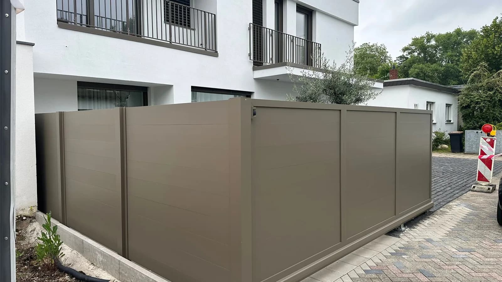 Braungrauer Zaun aus waagerechten Elementen entlang eines gepflasterten Wegs neben einem hellen Haus.