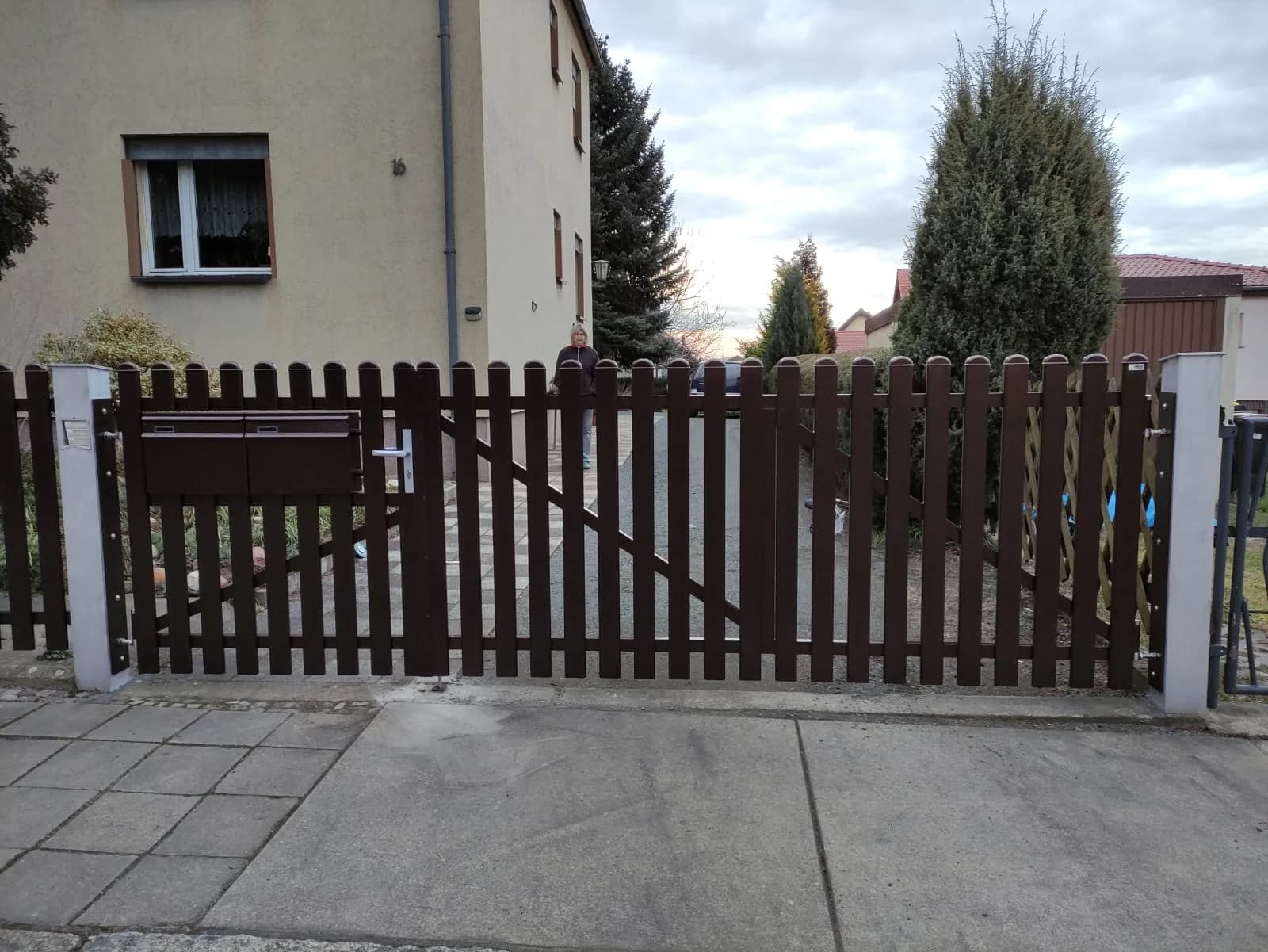 Braunes Tor mit senkrechten Stäben an einer gepflasterten Einfahrt vor einem Haus.