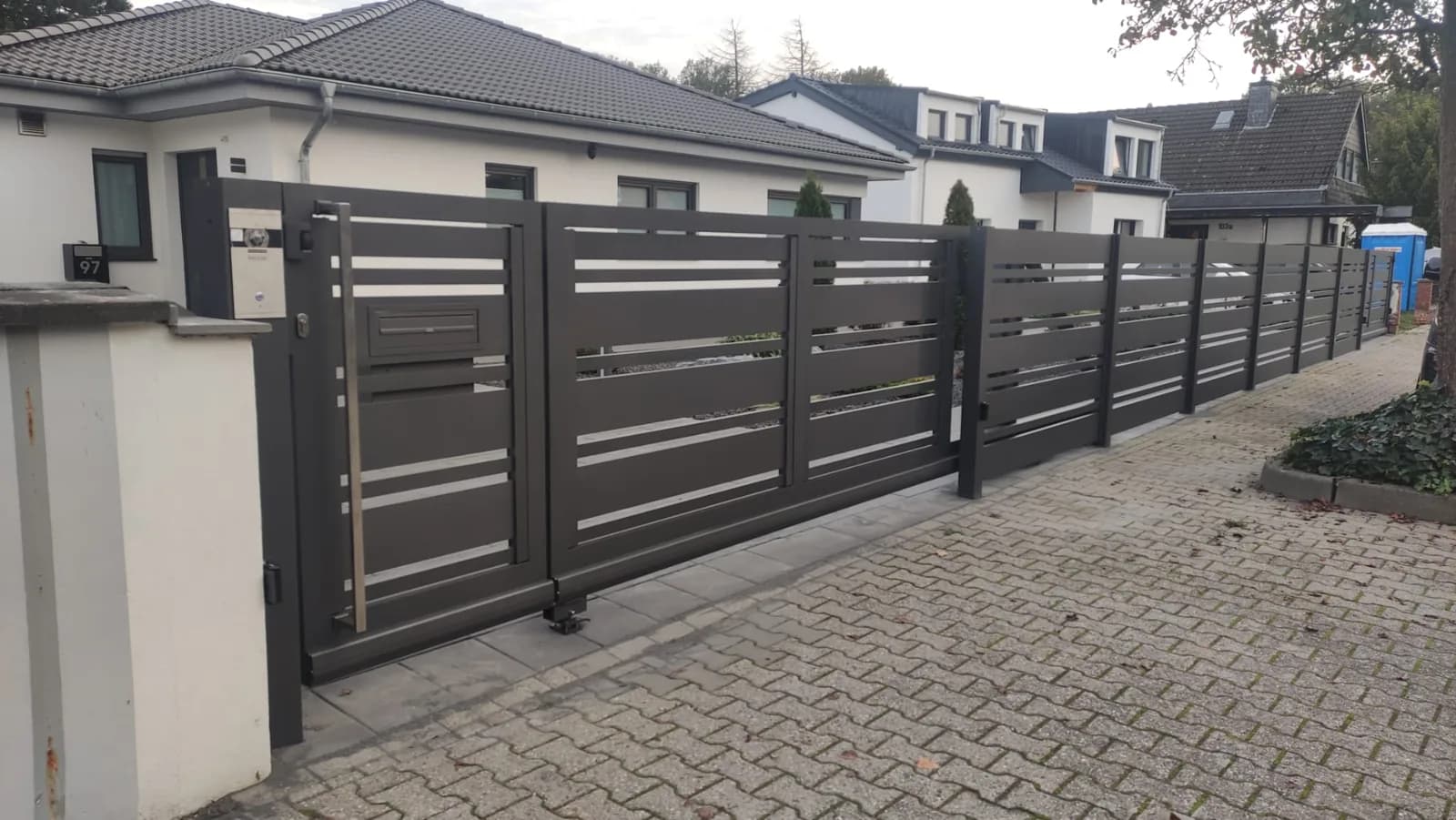 Langer dunkler Zaun aus waagerechten Lamellen mit hellen Pfosten an einer gepflasterten Einfahrt.