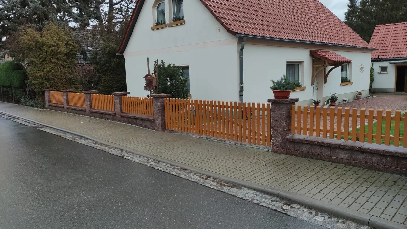 Brauner Zaun mit senkrechten Stäben und Tor entlang eines Gehwegs vor einem hellen Haus.