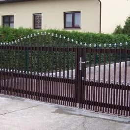 Braunes Tor mit senkrechten Stäben und hellen Zierspitzen, dahinter ein Haus.