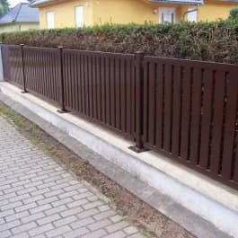Brauner Zaun mit senkrechten Stäben neben einer Hecke und einem Gehweg.