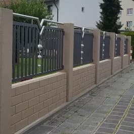 Dunkler Zaun mit senkrechten Stäben zwischen gemauerten Pfosten an einem Gehweg.