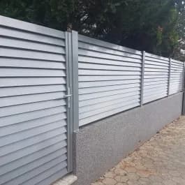 Hellgrauer Zaun aus waagerechten Elementen mit dunklen Pfosten auf einem Mauersockel.