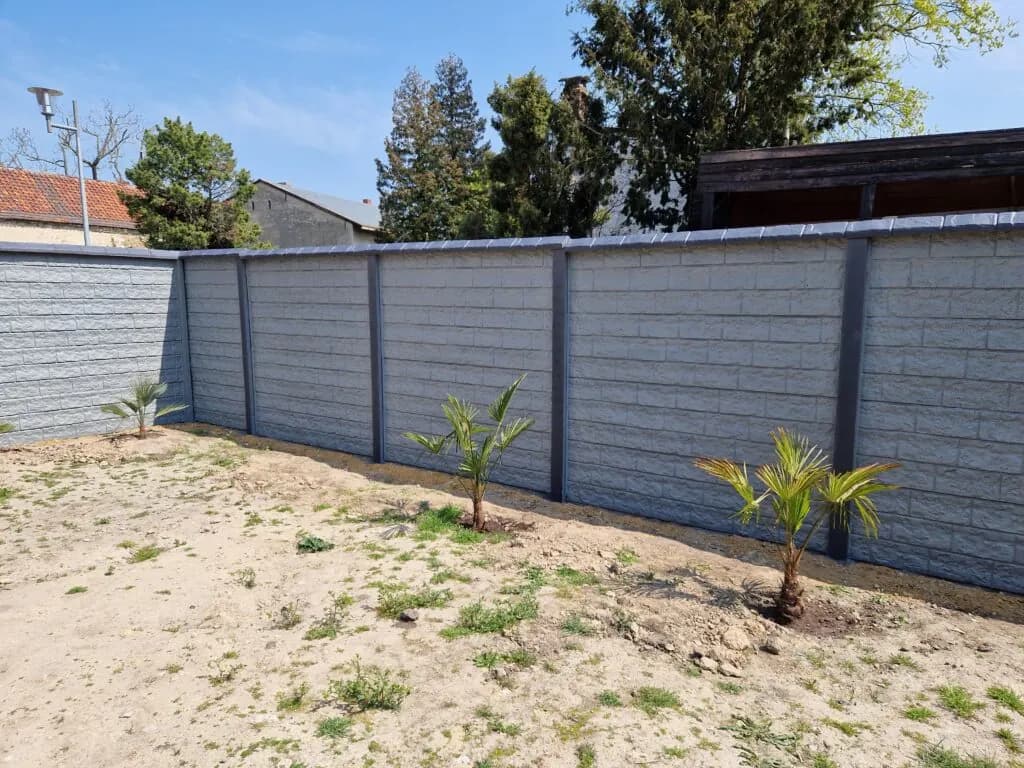 Gerader grauer Betonzaun mit dunklen Pfosten neben einer sandigen Gartenfläche mit kleinen Pflanzen.
