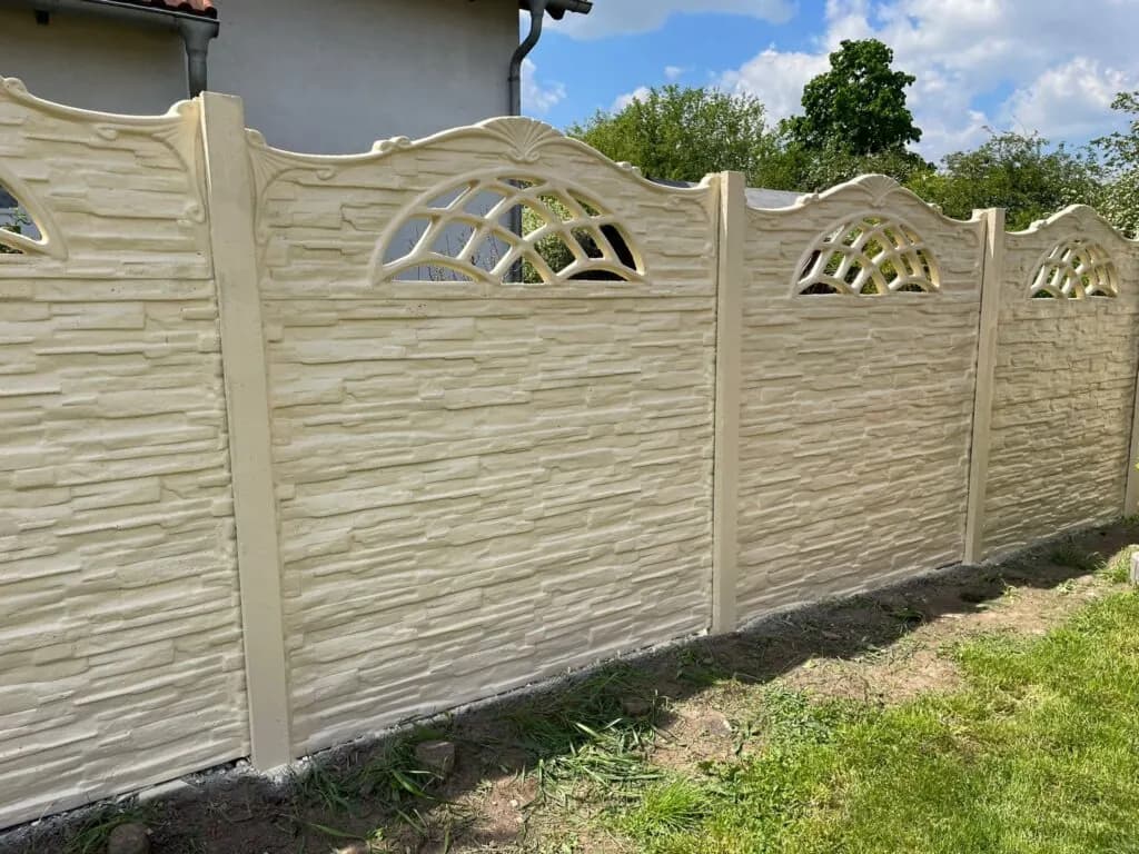 Heller Betonzaun mit Steinstruktur und bogenförmigen Öffnungen vor einer Hauswand.