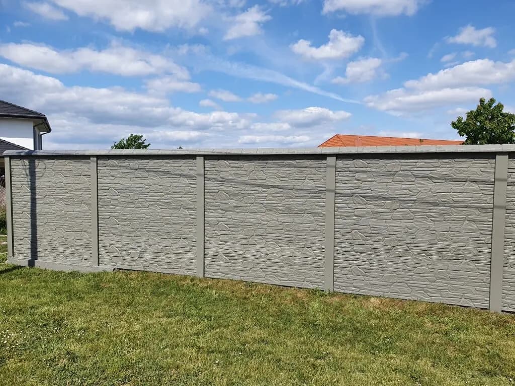 Langer hellgrauer Betonzaun mit waagerechter Struktur, dunklen Pfosten und Rasen im Vordergrund.