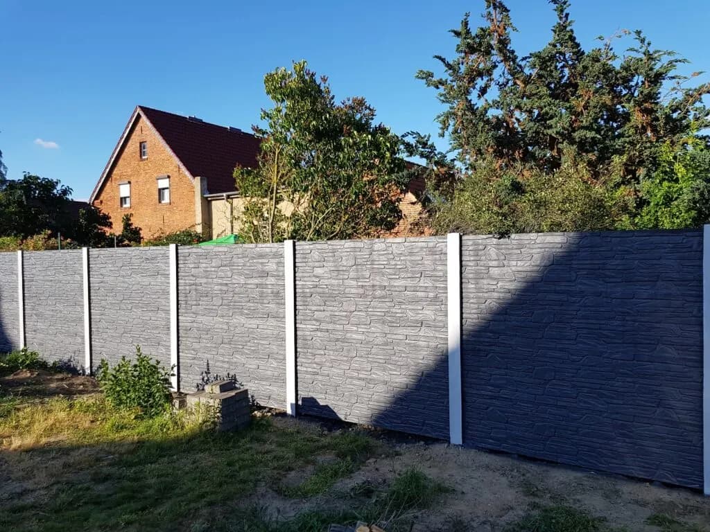 Gerader grauer Betonzaun mit hellen Pfosten und waagerechtem Relief vor einem Garten und Ziegelhaus.