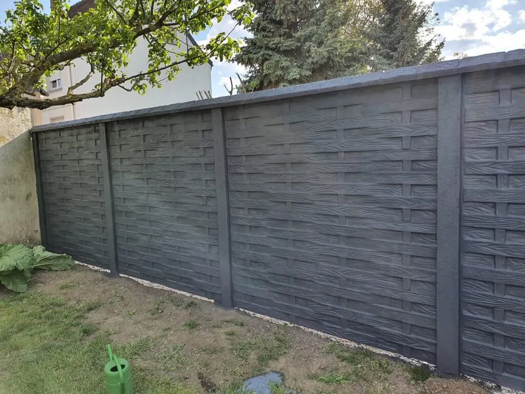 Dunkelgrauer Betonzaun mit waagerechter Flechtstruktur unter Bäumen entlang eines Gartens.