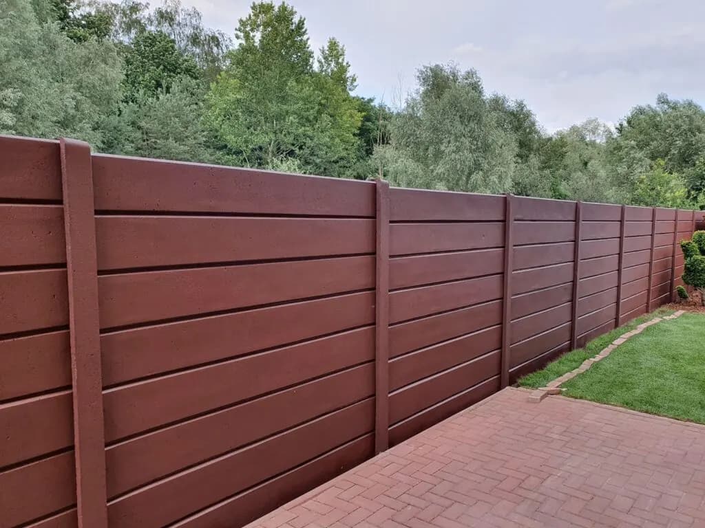 Brauner Betonzaun aus glatten waagerechten Elementen neben einem gepflasterten Weg und Rasen.