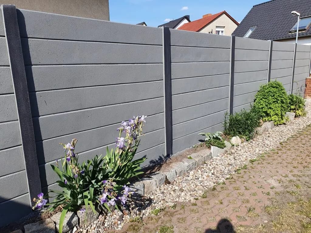 Dunkelgrauer Betonzaun aus glatten horizontalen Feldern neben einem Kiesbeet und Pflasterweg.