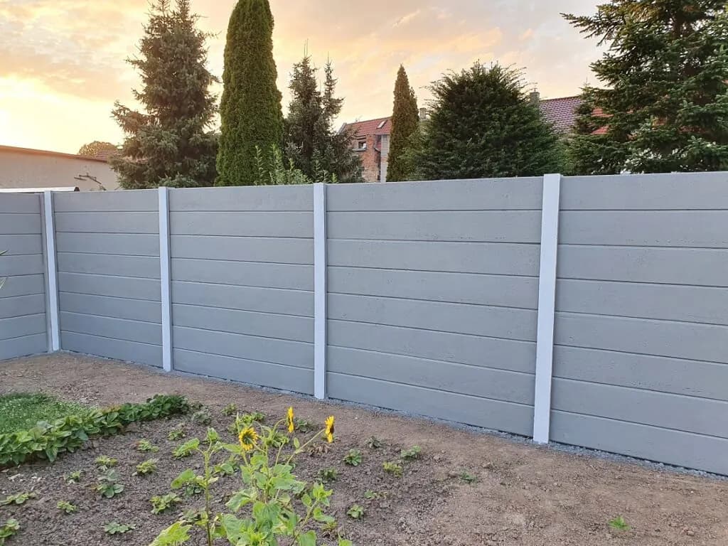 Hellgrauer Betonzaun mit waagerechten Platten und weißen Pfosten hinter einem bepflanzten Beet.
