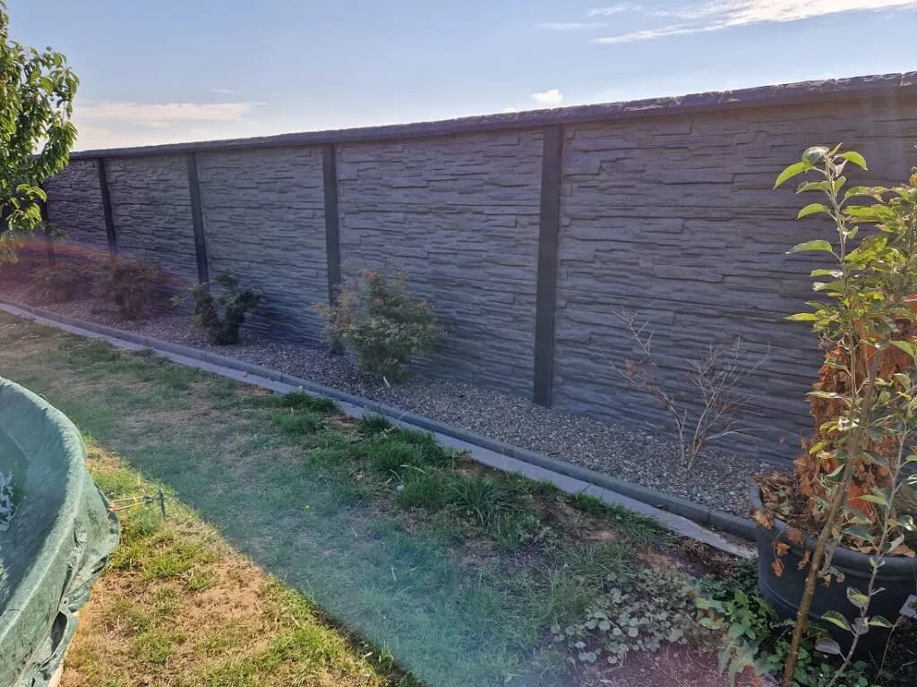 Dunkelgrauer Betonzaun mit Steinrelief und Abdeckungen entlang einer Rasenfläche mit jungen Sträuchern.
