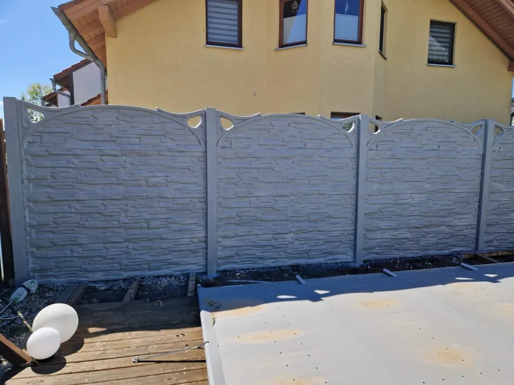 Grauer Betonzaun mit geschwungenen Oberkanten und runden Öffnungen vor einem gelben Haus.