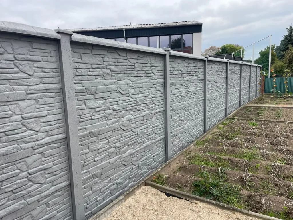 Langer hellgrauer Betonzaun mit Steinrelief und geraden Pfosten neben einem Gartenweg.