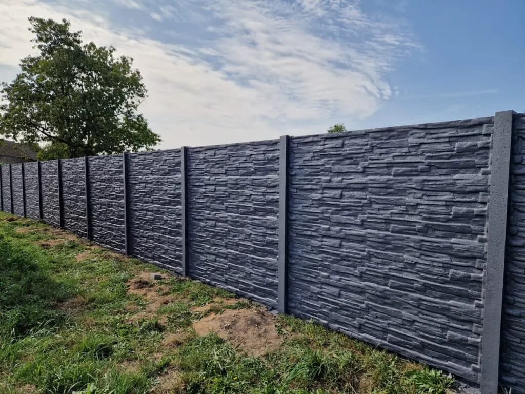 Dunkelgrauer Betonzaun mit Steinrelief entlang einer Rasenfläche und Bäumen im Hintergrund.