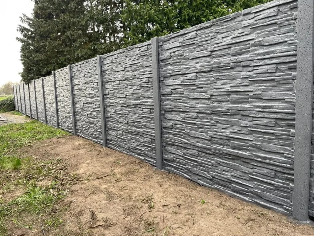 Hellgrauer Betonzaun mit horizontalem Steinrelief, gesehen entlang einer Grasfläche.