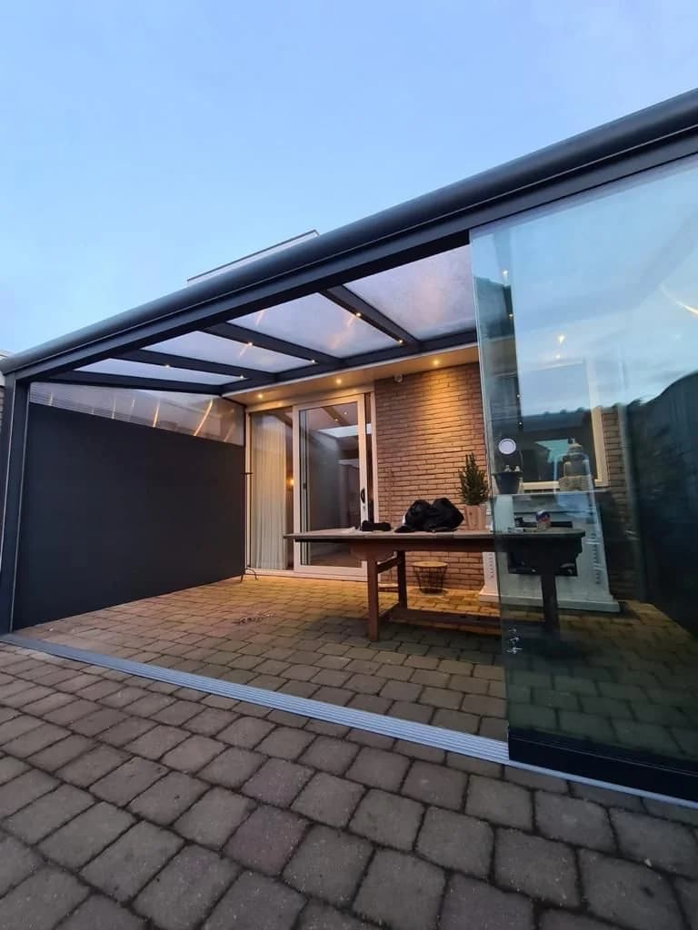 Überdachter Sitzbereich mit großflächigen Glaswänden an einem Haus, aufgenommen im Abendlicht.