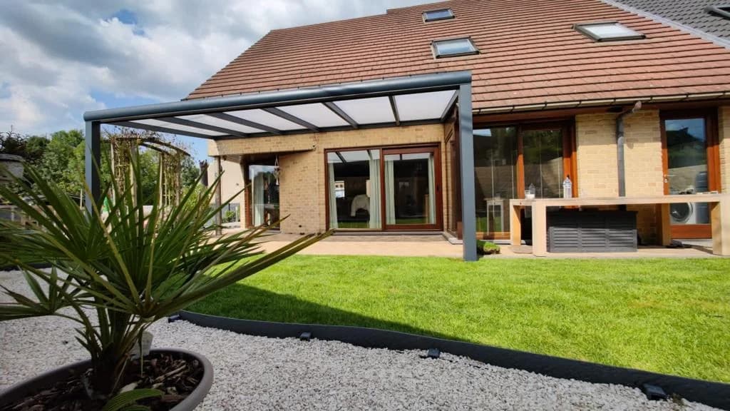 Offene Terrassenüberdachung mit dunklem Rahmen vor einem Haus, Garten und Rasenfläche im Vordergrund.