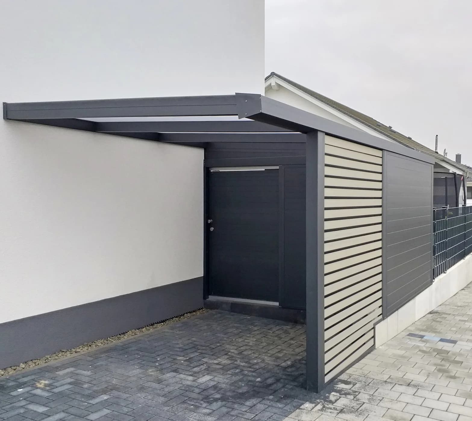 Überdachter Stellplatz mit seitlichen Lamellen neben einer weißen Hauswand und einer gepflasterten Zufahrt.