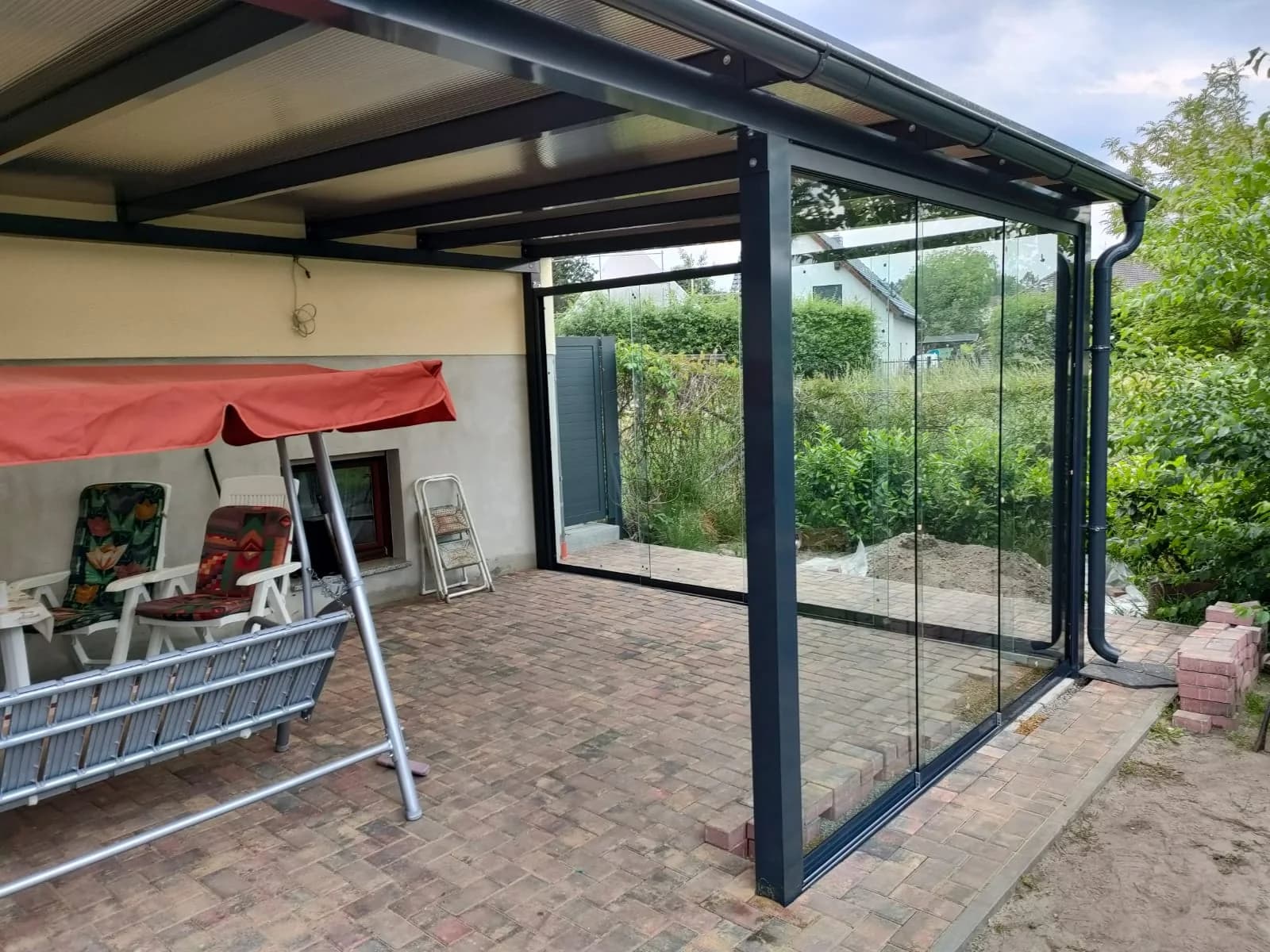 Überdachter Bereich mit großen seitlichen Glasscheiben, darunter Gartenmöbel und Leiter neben einer Hauswand.