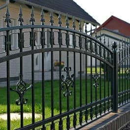 Dunkler Zierzaun mit gebogener Oberkante, Spitzen und Ornamenten vor einer Rasenfläche und Häusern.
