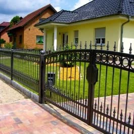 Dunkles Ziertor mit Spitzen und runden Elementen an einem gepflasterten Weg vor einem gelben Haus.