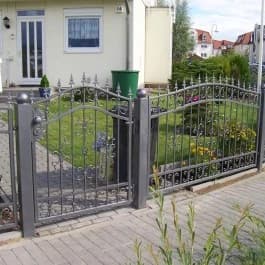 Graues Ziertor mit gebogener Oberkante und angrenzenden Zaunfeldern vor einem hellen Haus.
