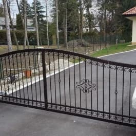 Dunkles zweiflügeliges Tor mit gebogener Oberkante und Ornamenten vor einer Einfahrt.