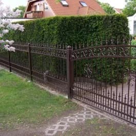 Dunkler Zierzaun und Tor entlang einer hohen Hecke und eines Gartenwegs.