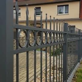 Nahansicht eines grauen Zauns mit Spitzen und ringförmigen Elementen an einer gepflasterten Einfahrt.