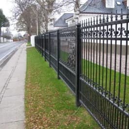 Langer dunkler Zierzaun mit Spitzen entlang einer Straße und eines Grünstreifens.