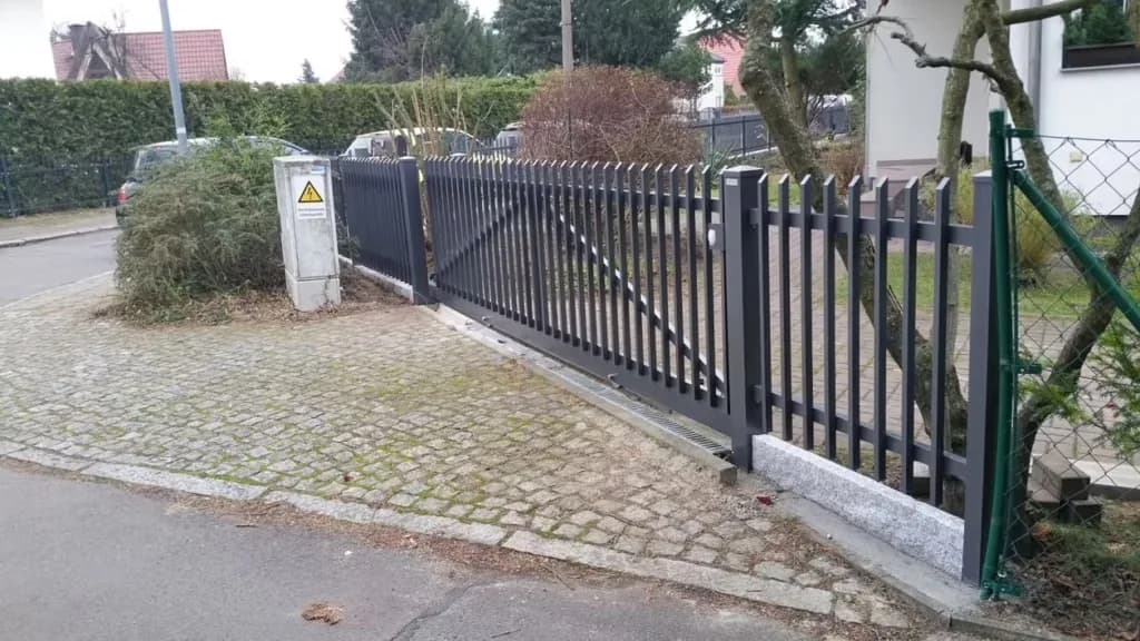 Dunkles Schiebetor mit senkrechten Stäben an einer gepflasterten Einfahrt und angrenzendem Zaun.