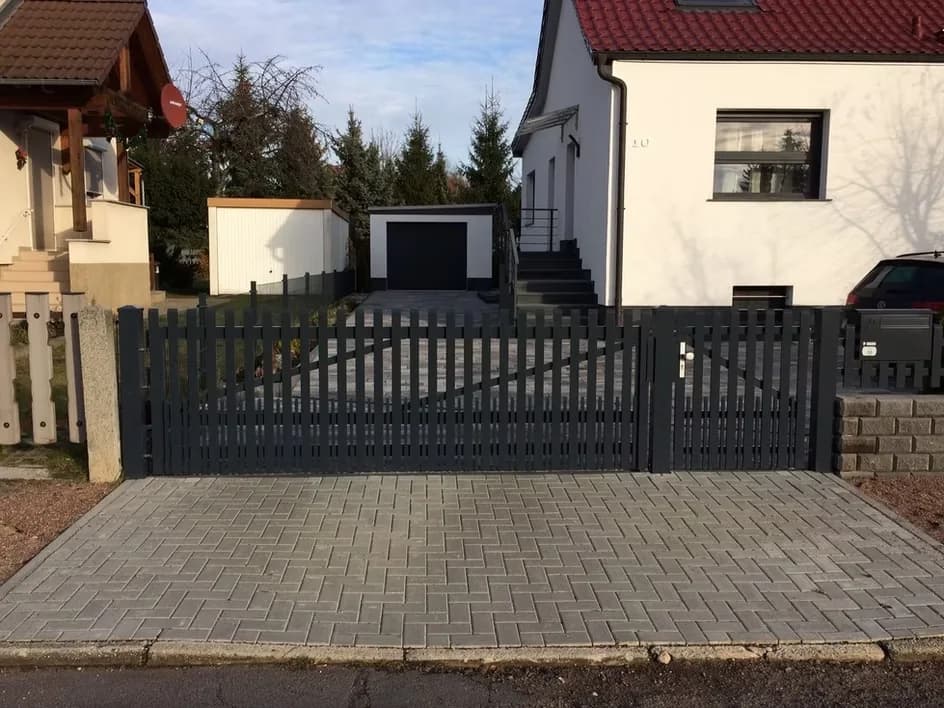 Dunkles Tor mit senkrechten Stäben und daneben ein schmaler Zugang vor einem Wohnhaus.