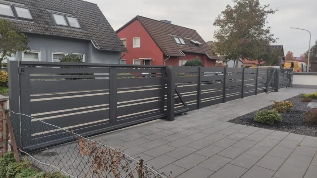 Dunkles Schiebetor mit waagerechten Streben vor einer langen passenden Zaunanlage an einer gepflasterten Einfahrt.