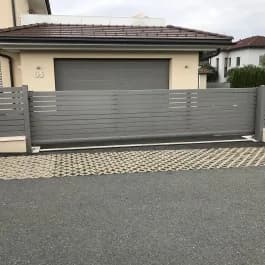 Graues Schiebetor mit waagerechten Streben vor einer Garage und einem gepflasterten Bereich.